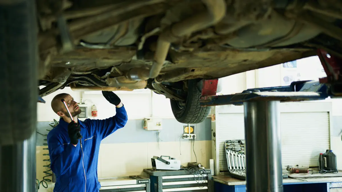 A mechanic inspecting the underside of a raised vehicle with a hand lamp.