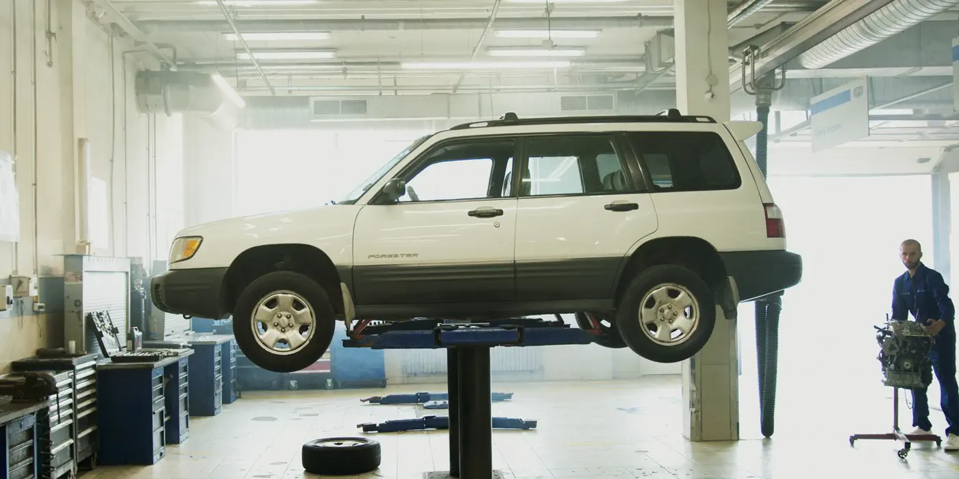 A vehicle raised on a workshop ramp with a mechanic working at the bench behind.